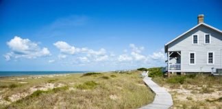 A long wooden walkway leading up to a modest beachfront house. The day is sunny with some cloud cover.