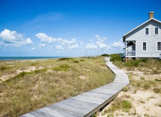 A long wooden walkway leading up to a modest beachfront house. The day is sunny with some cloud cover.