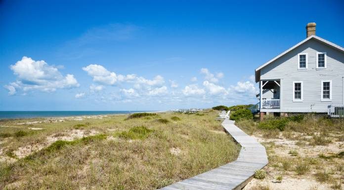 A long wooden walkway leading up to a modest beachfront house. The day is sunny with some cloud cover.