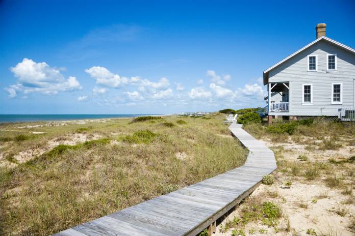 A long wooden walkway leading up to a modest beachfront house. The day is sunny with some cloud cover.