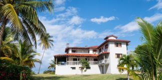 A Caribbean style home with white stone walls and a red tile roof sitting on a lawn with palm trees.