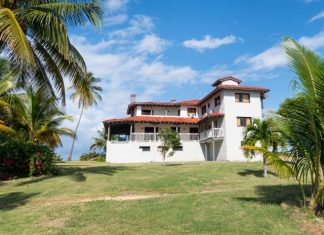 A Caribbean style home with white stone walls and a red tile roof sitting on a lawn with palm trees.