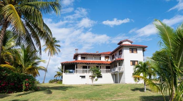 A Caribbean style home with white stone walls and a red tile roof sitting on a lawn with palm trees.
