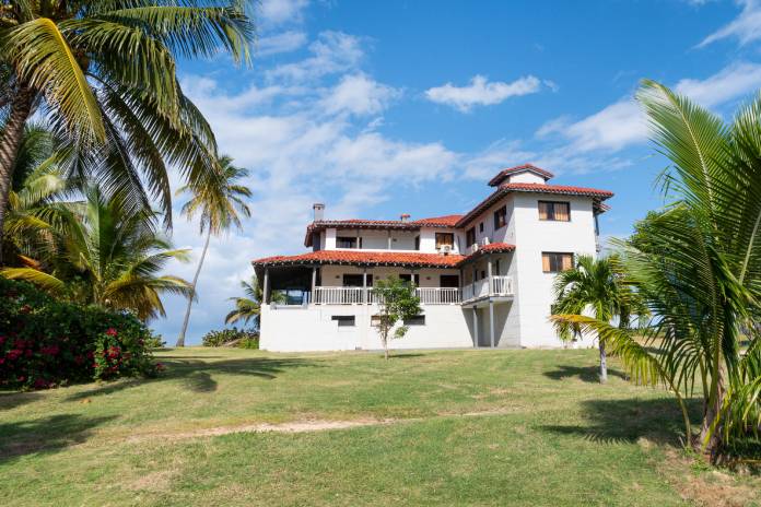 A Caribbean style home with white stone walls and a red tile roof sitting on a lawn with palm trees.