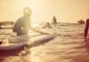 A group of people surfing in open waters beneath clear skies at sunrise. A gentle wave is coming toward them.