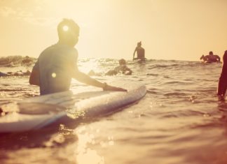 A group of people surfing in open waters beneath clear skies at sunrise. A gentle wave is coming toward them.