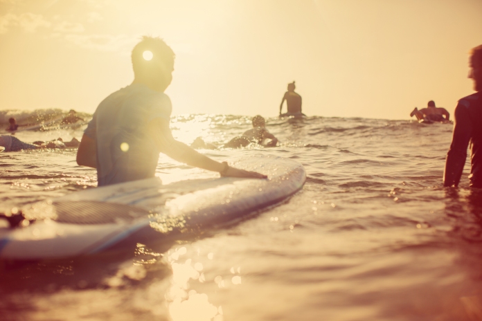 A group of people surfing in open waters beneath clear skies at sunrise. A gentle wave is coming toward them.