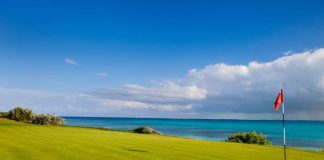 A golf course green overlooks the crystal-clear, turquoise ocean. A red golf pin flag sits in the hole.