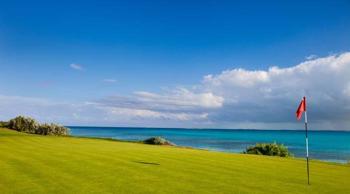A golf course green overlooks the crystal-clear, turquoise ocean. A red golf pin flag sits in the hole.