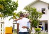 A couple embracing each other and holding the keys to their new home in the Caribbean, which is visible behind them.