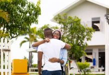 A couple embracing each other and holding the keys to their new home in the Caribbean, which is visible behind them.