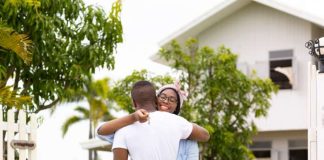 A couple embracing each other and holding the keys to their new home in the Caribbean, which is visible behind them.