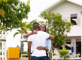 A couple embracing each other and holding the keys to their new home in the Caribbean, which is visible behind them.