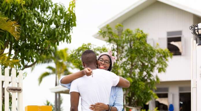 A couple embracing each other and holding the keys to their new home in the Caribbean, which is visible behind them.