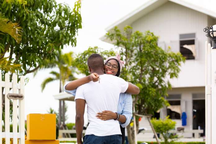 A couple embracing each other and holding the keys to their new home in the Caribbean, which is visible behind them.