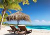 Two wooden lounge chairs sit under a straw umbrella on a sandy white beach. The sun shines on the beach and clear ocean water.