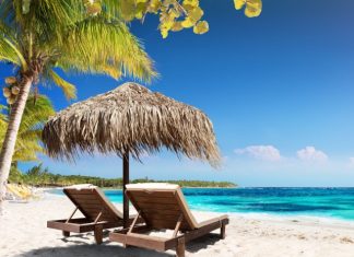 Two wooden lounge chairs sit under a straw umbrella on a sandy white beach. The sun shines on the beach and clear ocean water.