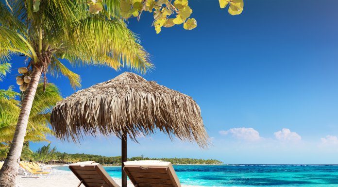 Two wooden lounge chairs sit under a straw umbrella on a sandy white beach. The sun shines on the beach and clear ocean water.