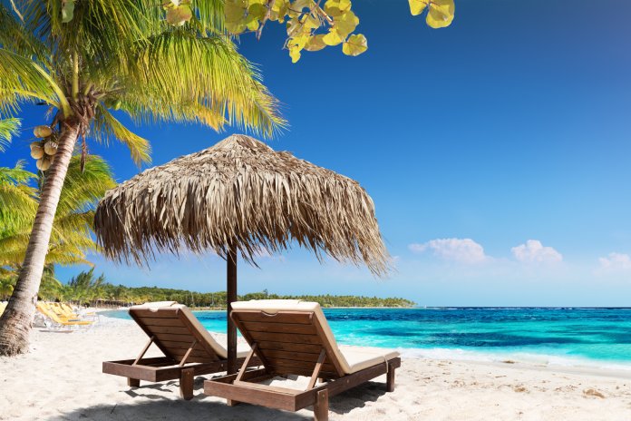 Two wooden lounge chairs sit under a straw umbrella on a sandy white beach. The sun shines on the beach and clear ocean water.