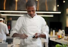 An African American chef sharpening a knife in a restaurant kitchen. Other chefs are in the background.