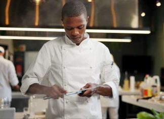 An African American chef sharpening a knife in a restaurant kitchen. Other chefs are in the background.