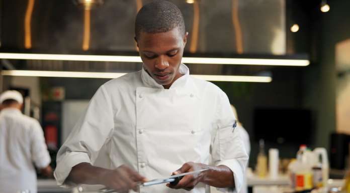 An African American chef sharpening a knife in a restaurant kitchen. Other chefs are in the background.