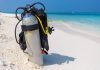 A silver scuba diving tank and breathing gear sit alone on a white-sand beach that overlooks a clear blue ocean.