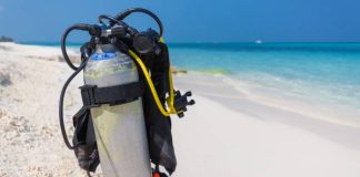 A silver scuba diving tank and breathing gear sit alone on a white-sand beach that overlooks a clear blue ocean.