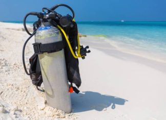 A silver scuba diving tank and breathing gear sit alone on a white-sand beach that overlooks a clear blue ocean.