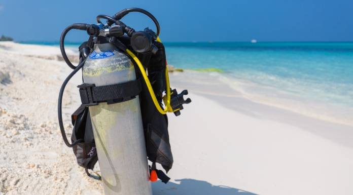 A silver scuba diving tank and breathing gear sit alone on a white-sand beach that overlooks a clear blue ocean.