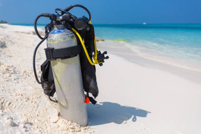 A silver scuba diving tank and breathing gear sit alone on a white-sand beach that overlooks a clear blue ocean.