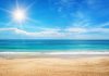 The crystal clear blue water splashes onto the white sandy beach. The sun shines bright in the blue sky.