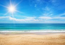 The crystal clear blue water splashes onto the white sandy beach. The sun shines bright in the blue sky.