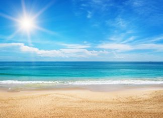 The crystal clear blue water splashes onto the white sandy beach. The sun shines bright in the blue sky.