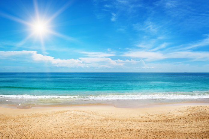 The crystal clear blue water splashes onto the white sandy beach. The sun shines bright in the blue sky.