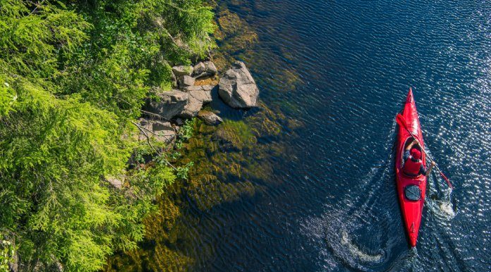 A person wearing an orange cap in an orange kayak on a blue lake paddles by vibrant green foliage and a rocky lake floor.