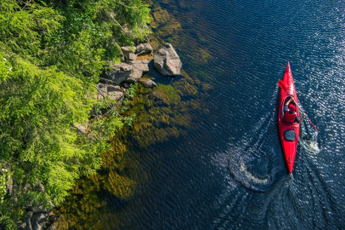 A person wearing an orange cap in an orange kayak on a blue lake paddles by vibrant green foliage and a rocky lake floor.