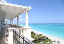 A balcony with a view of the beach and the ocean. The balcony is white, and there are chairs and tables on it.