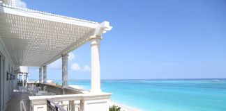 A balcony with a view of the beach and the ocean. The balcony is white, and there are chairs and tables on it.