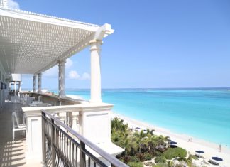 A balcony with a view of the beach and the ocean. The balcony is white, and there are chairs and tables on it.