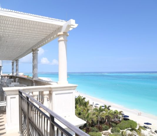 A balcony with a view of the beach and the ocean. The balcony is white, and there are chairs and tables on it.