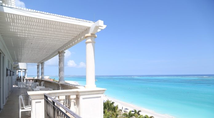 A balcony with a view of the beach and the ocean. The balcony is white, and there are chairs and tables on it.