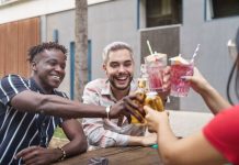 Three friends smile as they sit at a wooden picnic table outside. They clink their cocktails and beer bottles together.