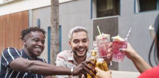 Three friends smile as they sit at a wooden picnic table outside. They clink their cocktails and beer bottles together.