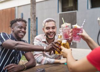 Three friends smile as they sit at a wooden picnic table outside. They clink their cocktails and beer bottles together.