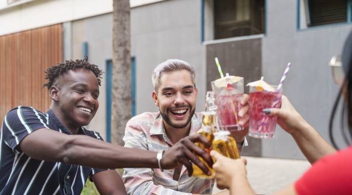 Three friends smile as they sit at a wooden picnic table outside. They clink their cocktails and beer bottles together.