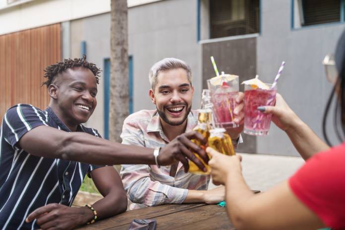 Three friends smile as they sit at a wooden picnic table outside. They clink their cocktails and beer bottles together.