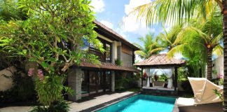 A villa with a pool, lounging chairs, and palm trees. It's a sunny day with a few clouds in the sky.