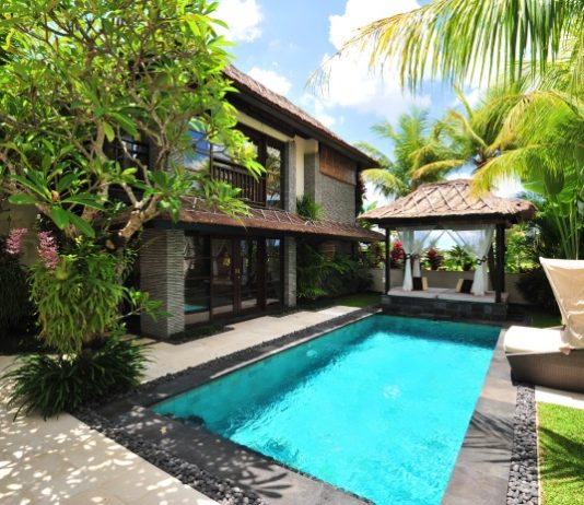 A villa with a pool, lounging chairs, and palm trees. It's a sunny day with a few clouds in the sky.