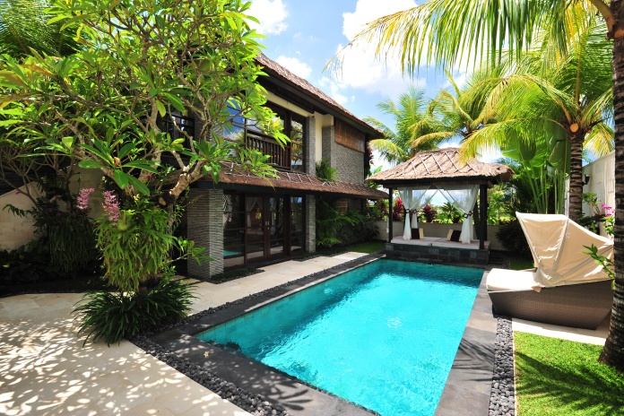 A villa with a pool, lounging chairs, and palm trees. It's a sunny day with a few clouds in the sky.